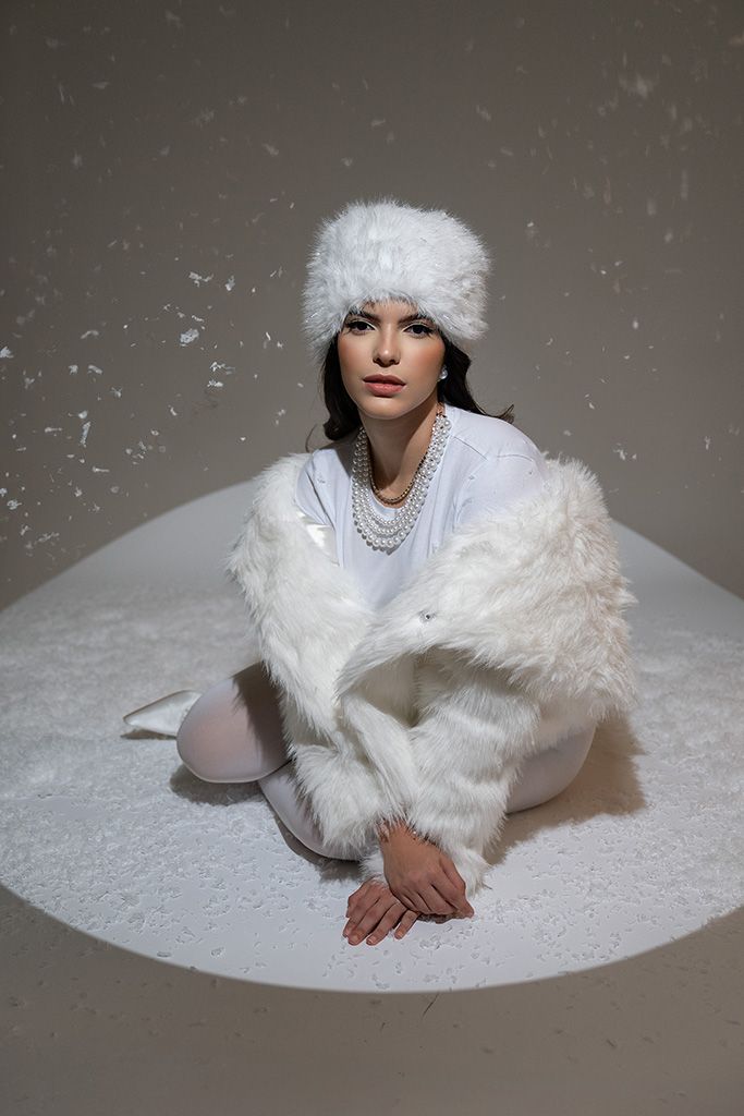 Woman in white fur coat and hat sitting gracefully with pearl necklace, surrounded by falling snow in studio.