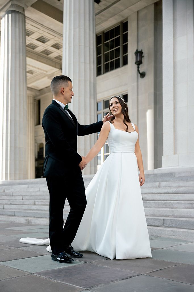 Bride and groom hold hands on grand stone steps with columns, she in white gown, he in black tuxedo.