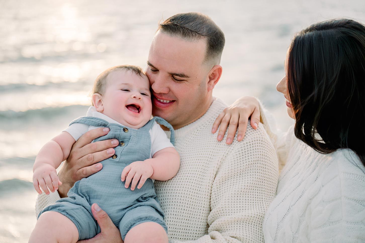 A laughing baby in blue romper held by smiling parents on a beach at sunset.
