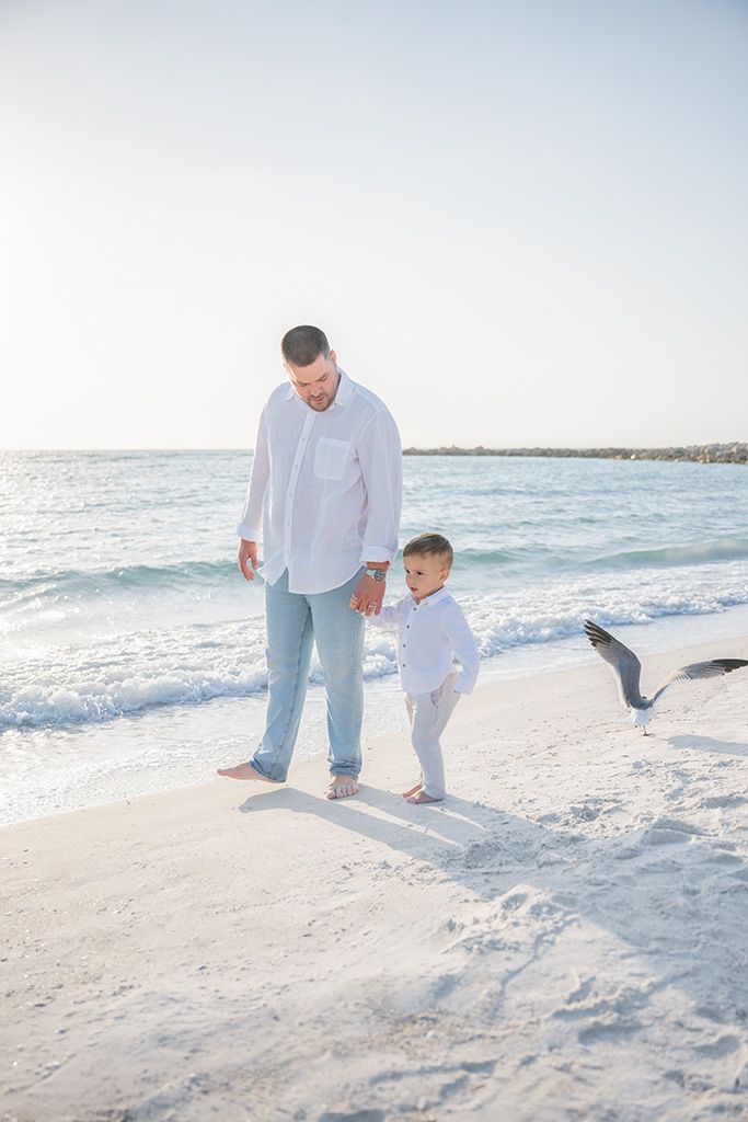 Father and young son holding hands walking barefoot on a sandy beach.