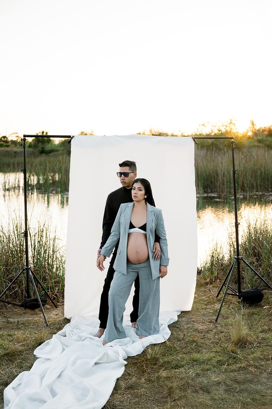Pregnant woman in gray suit poses with man in black outdoors near lake at sunset with white photo backdrop.