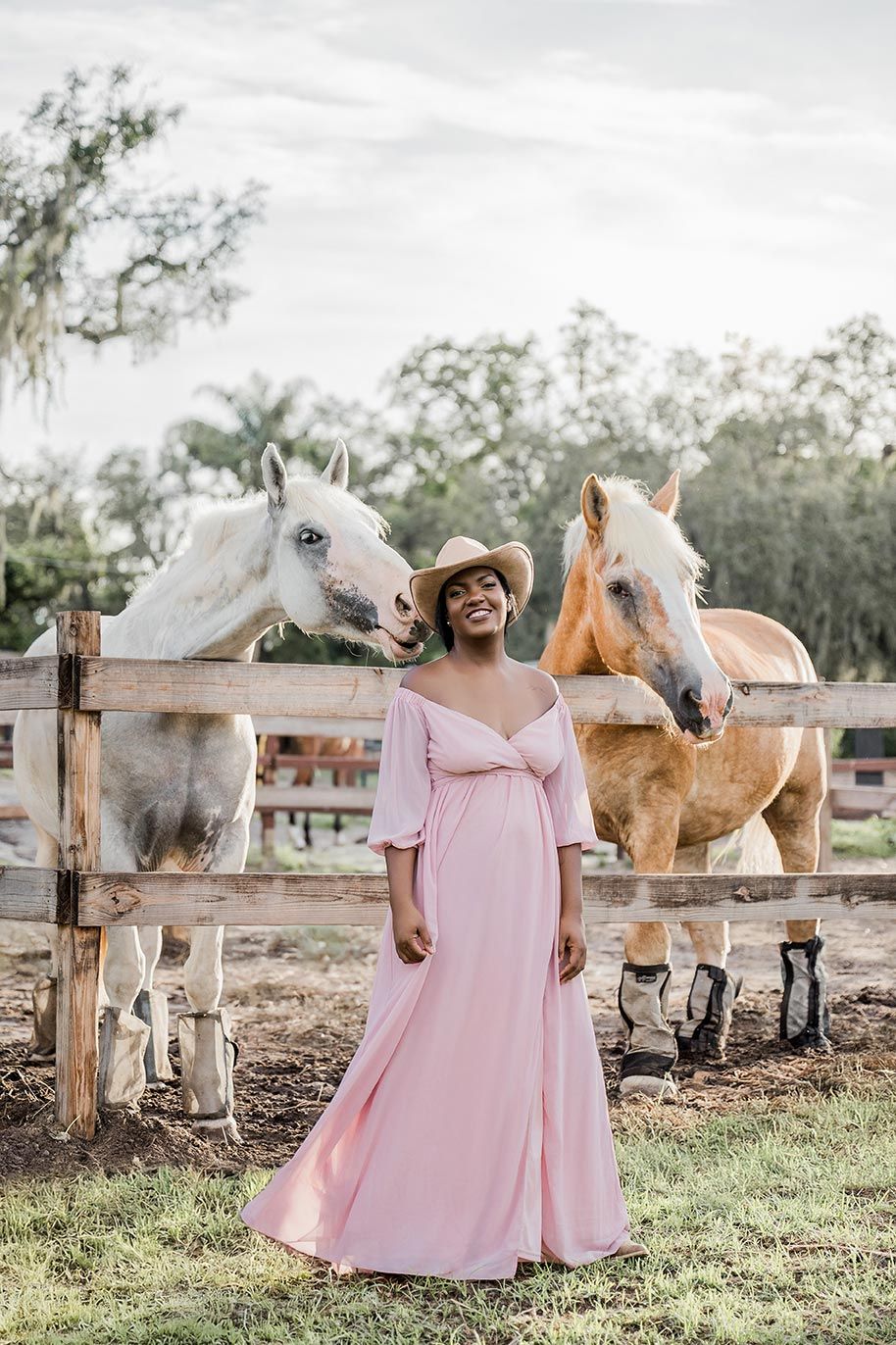 A person in a flowing pink dress interacts with white and palomino horses in a rustic fenced pasture.