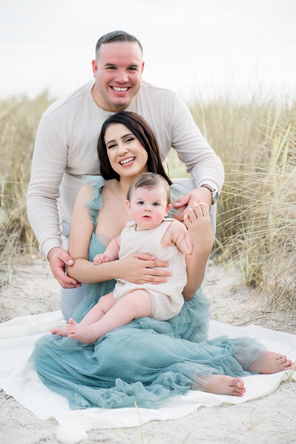 Family wearing white and turquoise sits together in beach grass during a coastal photoshoot.