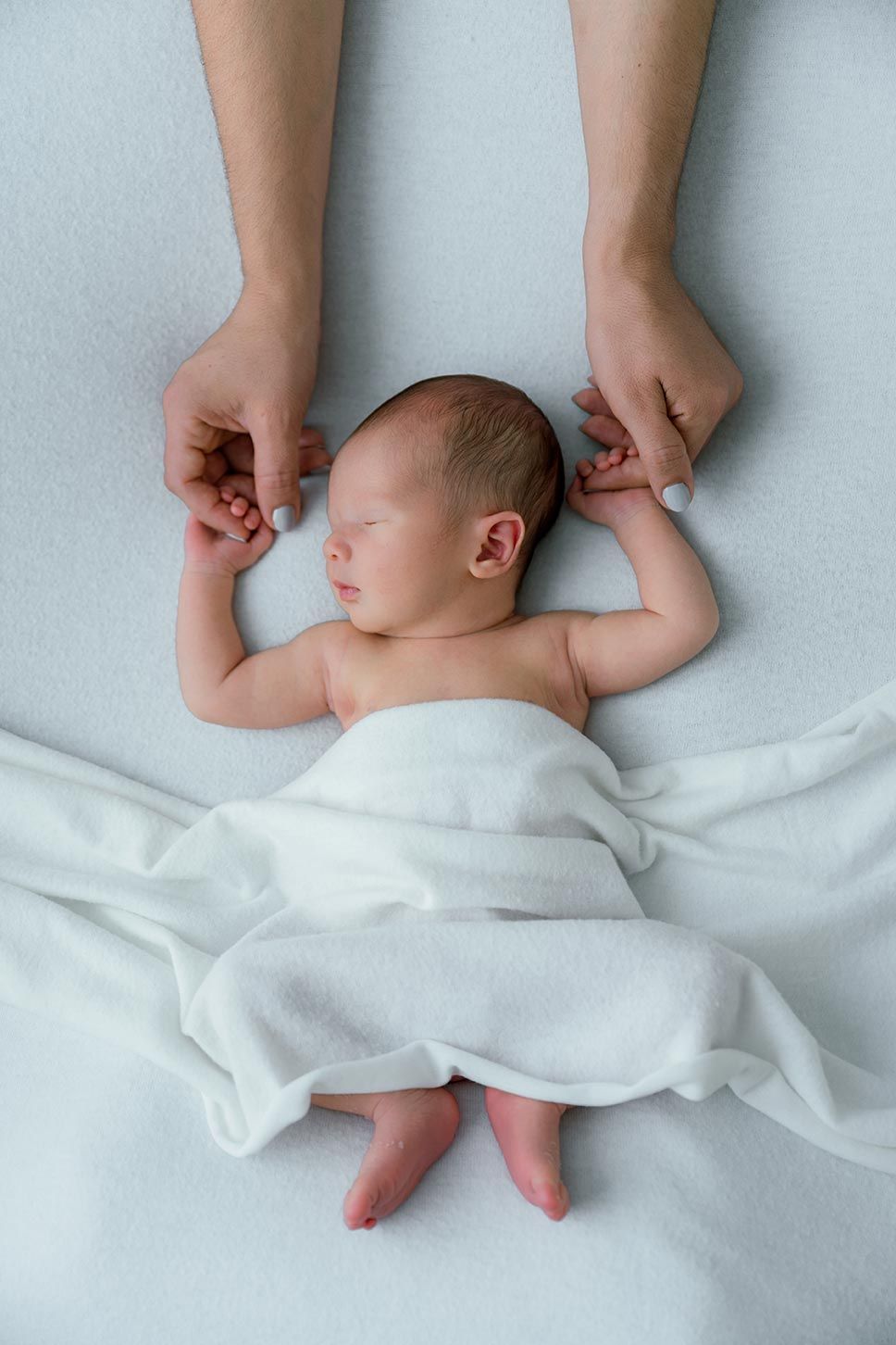 A sleeping newborn baby lies on white bedding while gentle hands hold their tiny fingers.