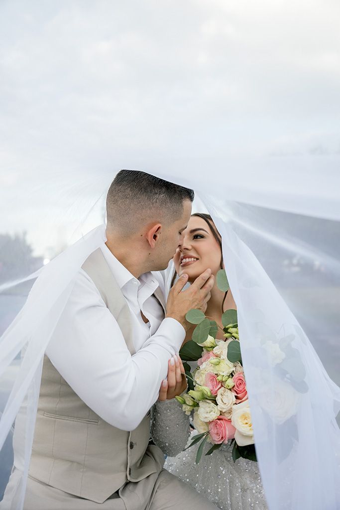 Romantic wedding couple almost kissing under billowing white veil outdoors, bride holding elegant floral bouquet.