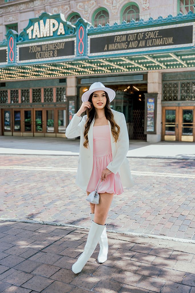 Woman in pink dress, white blazer, hat, and boots posing outside Tampa Theatre marquee.
