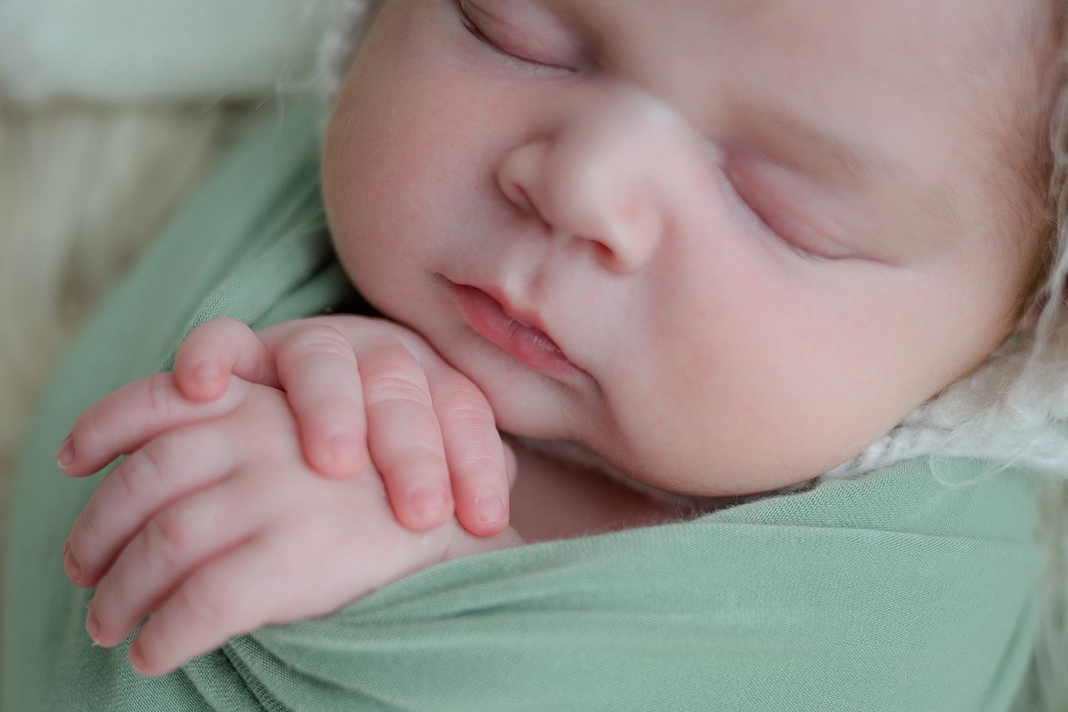 Peaceful newborn baby sleeping wrapped in a soft green swaddle blanket with tiny hands folded together.