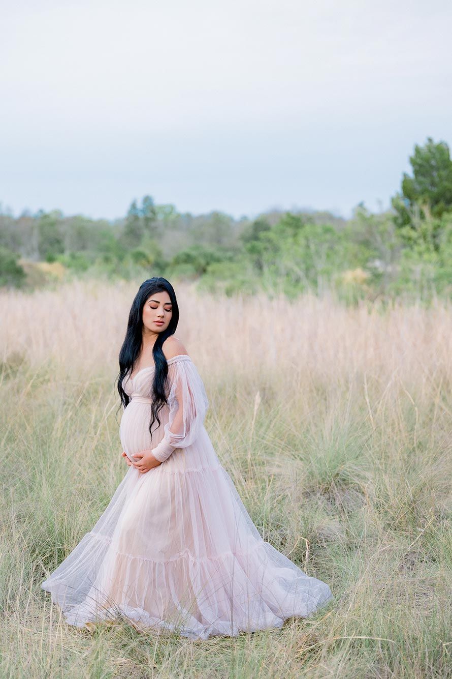 Pregnant woman in blush pink tulle gown standing in golden grass field during outdoor maternity photoshoot.