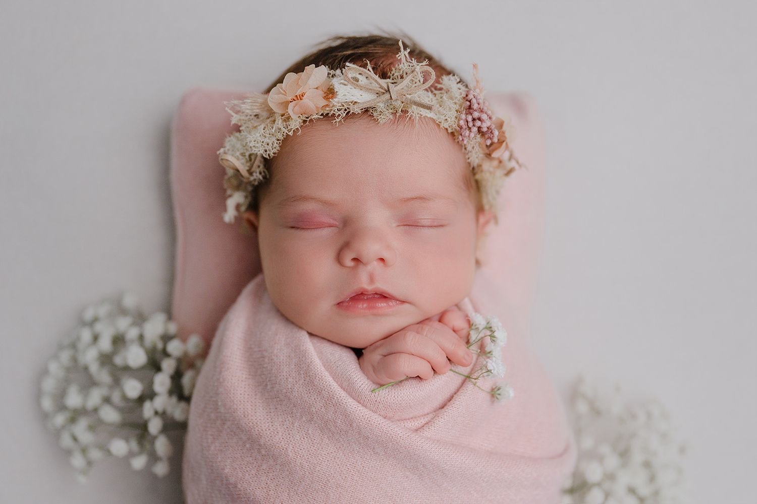 Sleeping newborn baby girl wrapped in pink swaddle with floral crown headband on white background.