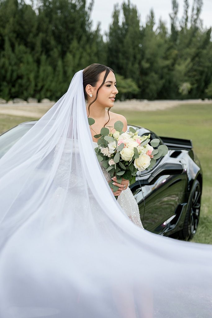 Bride in lace gown holds white floral bouquet, veil flowing, standing beside black car with green trees backdrop.