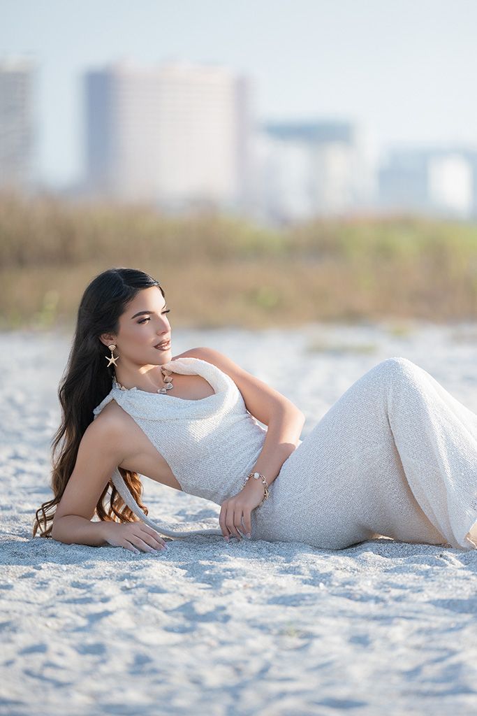 Woman in white gown posing on sandy beach with city skyline in background.