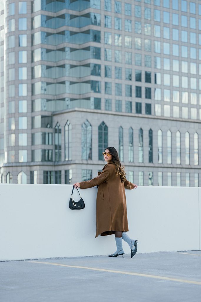 Woman in brown coat holding black bag poses on urban rooftop with modern glass skyscraper backdrop.