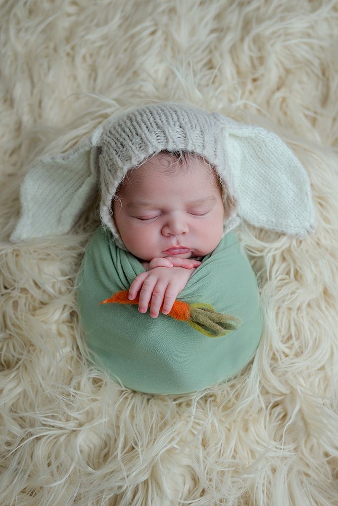 Sleeping newborn baby wrapped in green swaddle, wearing knit bunny ear bonnet, holding a tiny carrot on fluffy cream rug.