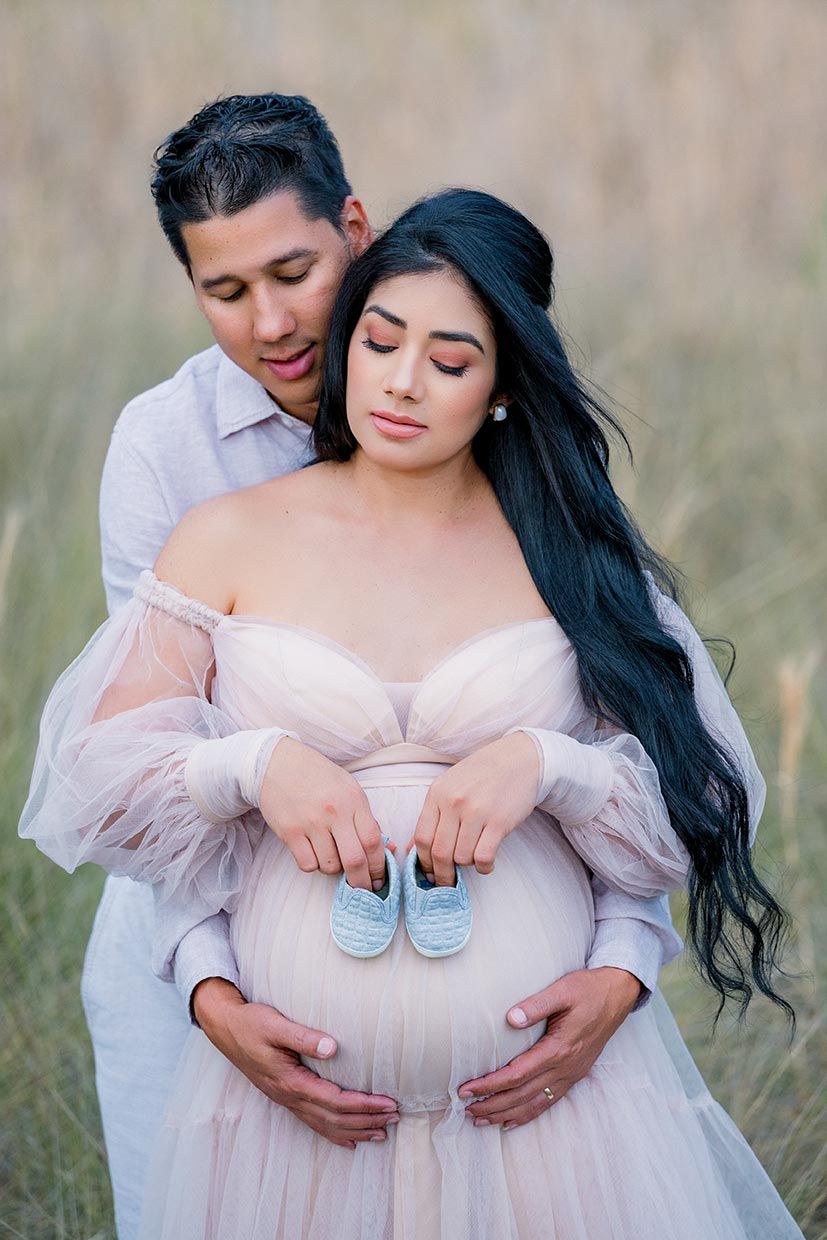 Couple in a field holding tiny blue baby shoes on pregnant belly, wearing a blush tulle gown for maternity photos.
