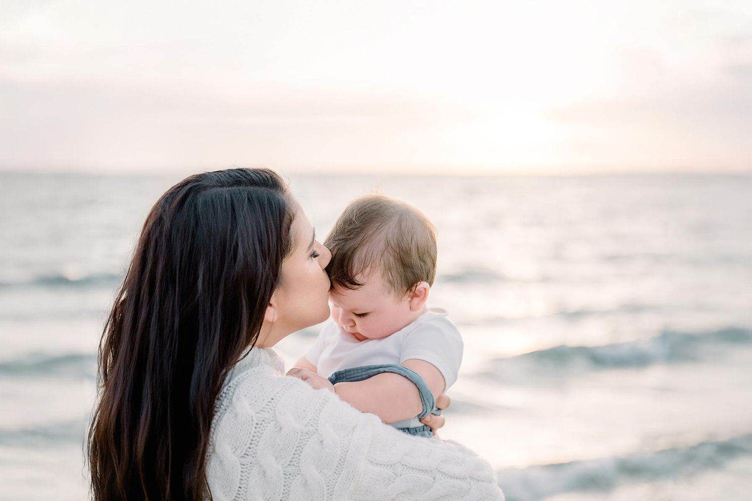 A mother tenderly kisses her baby by the ocean at sunset, both dressed in white.