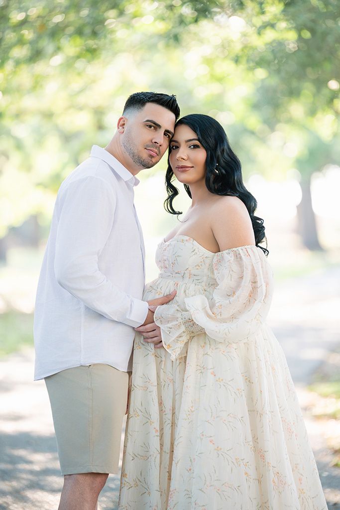 Couple poses outdoors, man in white shirt and woman in floral off-shoulder gown, maternity photo session.