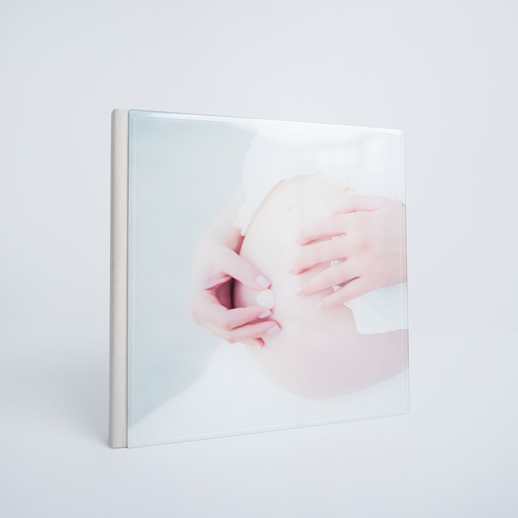 A square photo book featuring a soft, delicate image of hands gently cradling a pregnant belly on a white background.