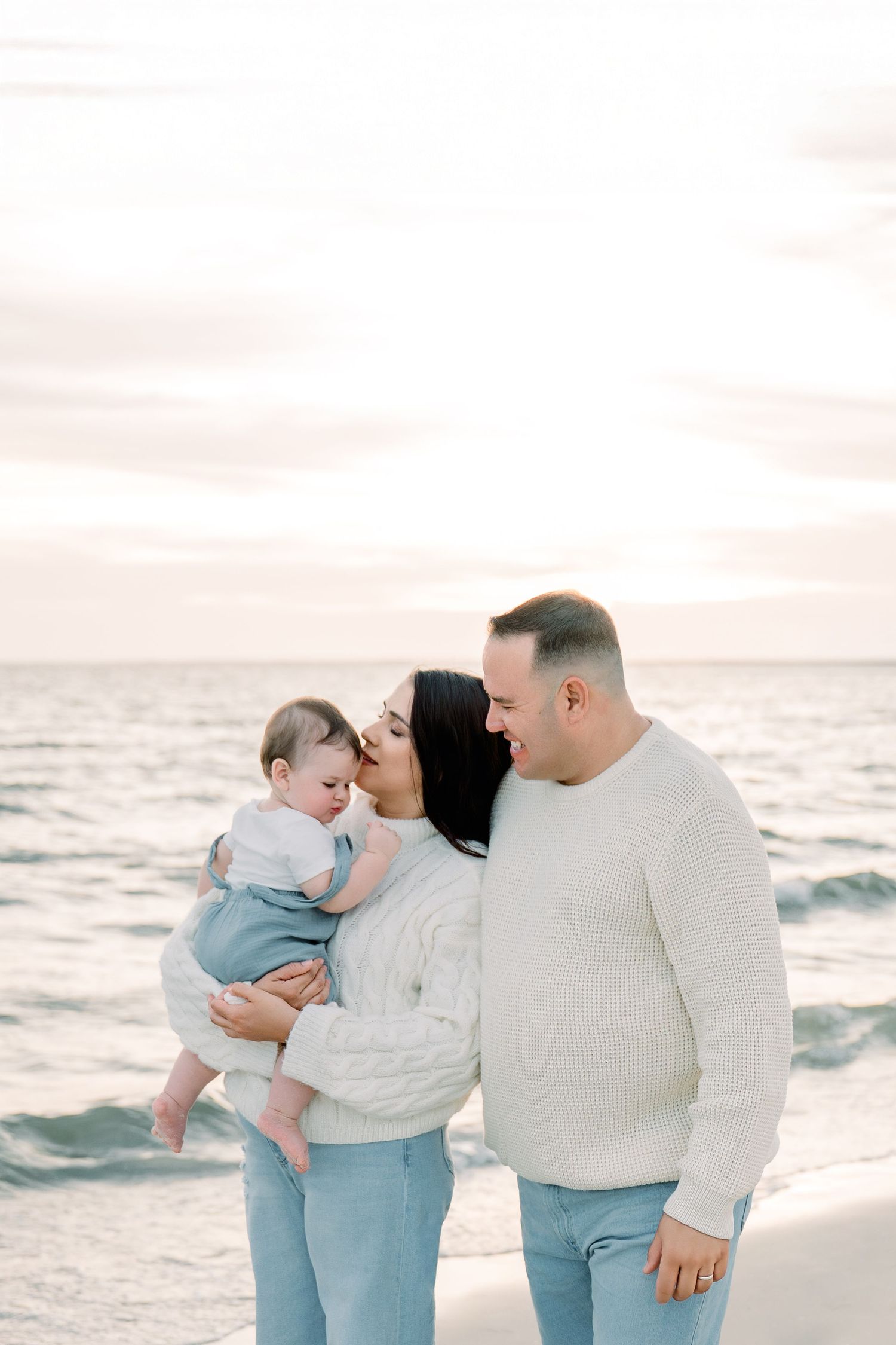 A family enjoys a tender moment together on a serene beach at sunset with ocean waves in the background.