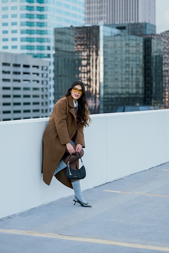 Stylish woman in camel coat and yellow sunglasses leans against white wall on rooftop holding black handbag.