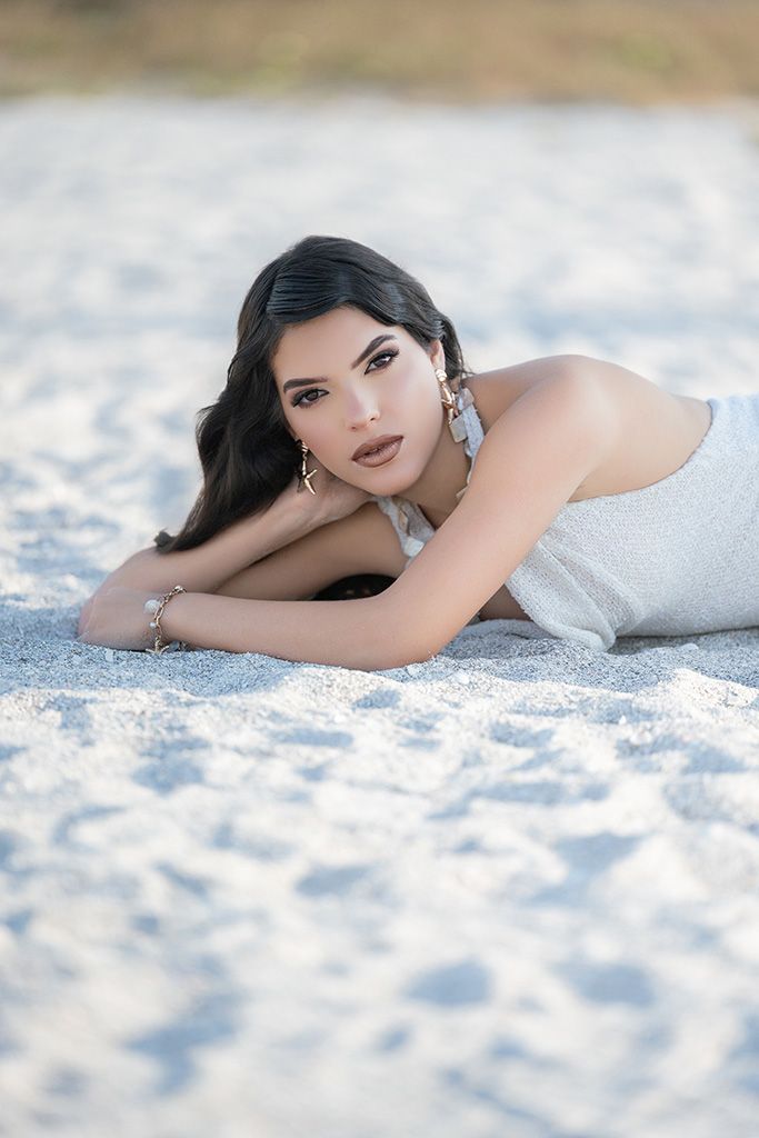 Young woman with dark hair lying on white sand beach, wearing silver outfit and elegant earrings, gazing at camera.