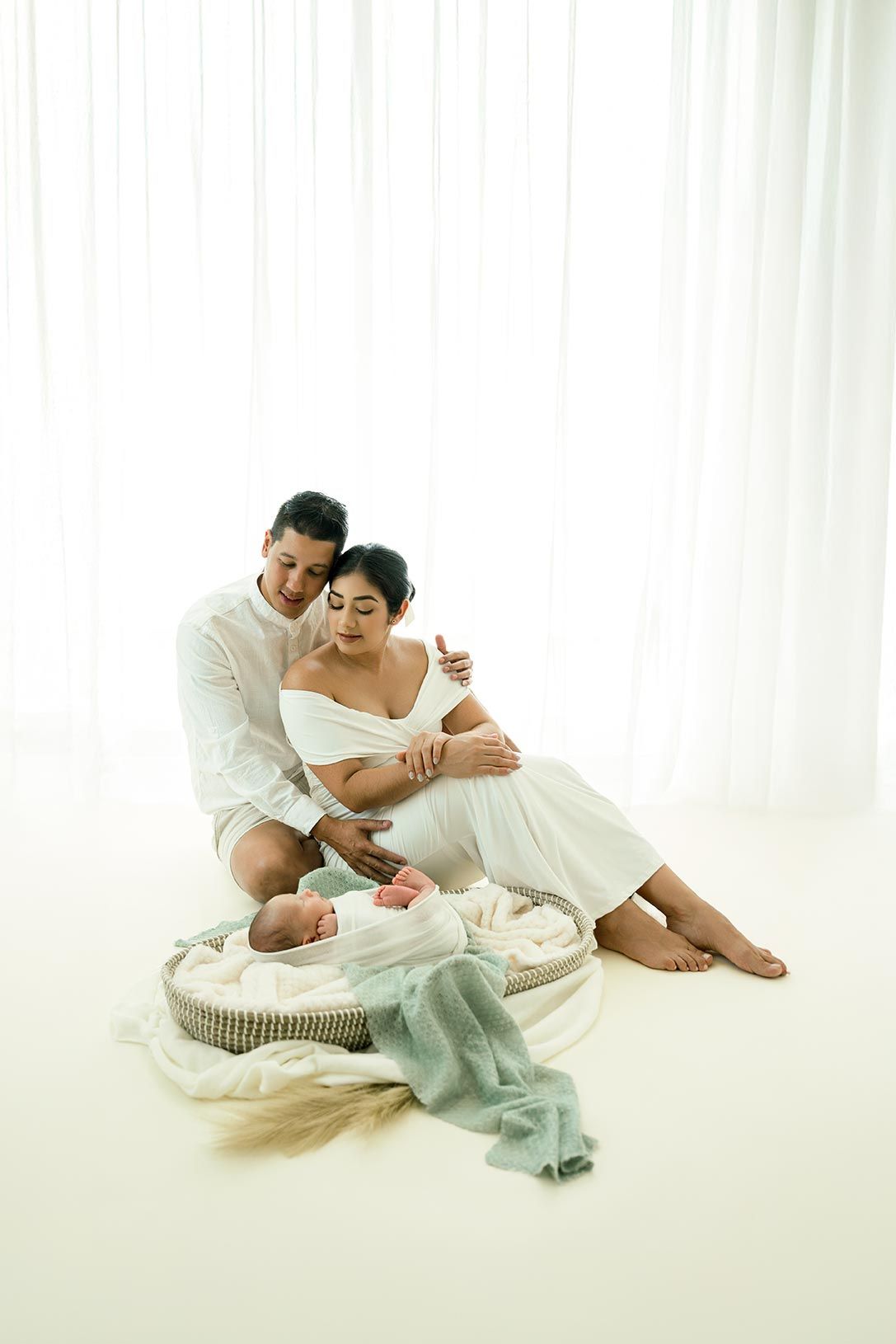 A couple relaxes together on the floor with their newborn baby in a bright, minimalist white studio setting.