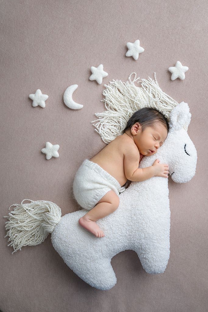 Newborn baby sleeping on a white unicorn stuffed animal surrounded by star and moon decorations.