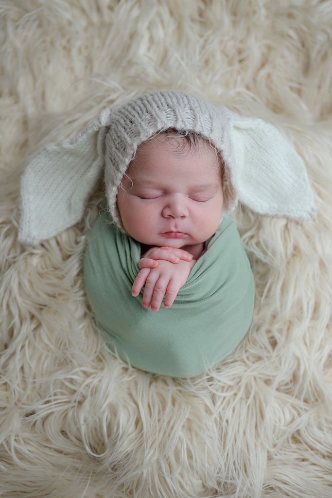 Newborn baby sleeping in green swaddle wrap wearing a knit bunny ear hat on fluffy cream rug.