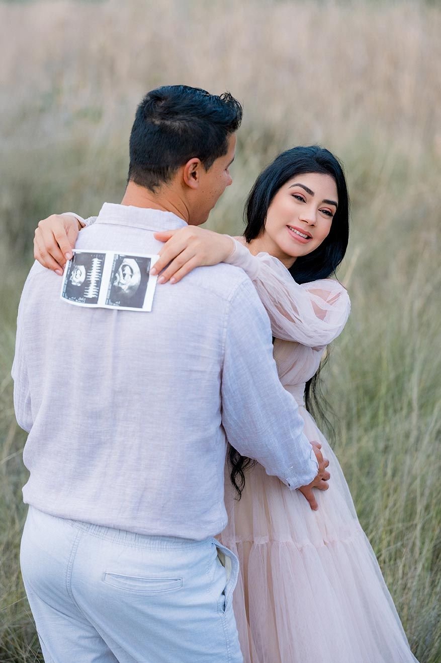 Couple embracing in a field, woman in pink dress smiling, man holding ultrasound photos, pregnancy announcement outdoors.