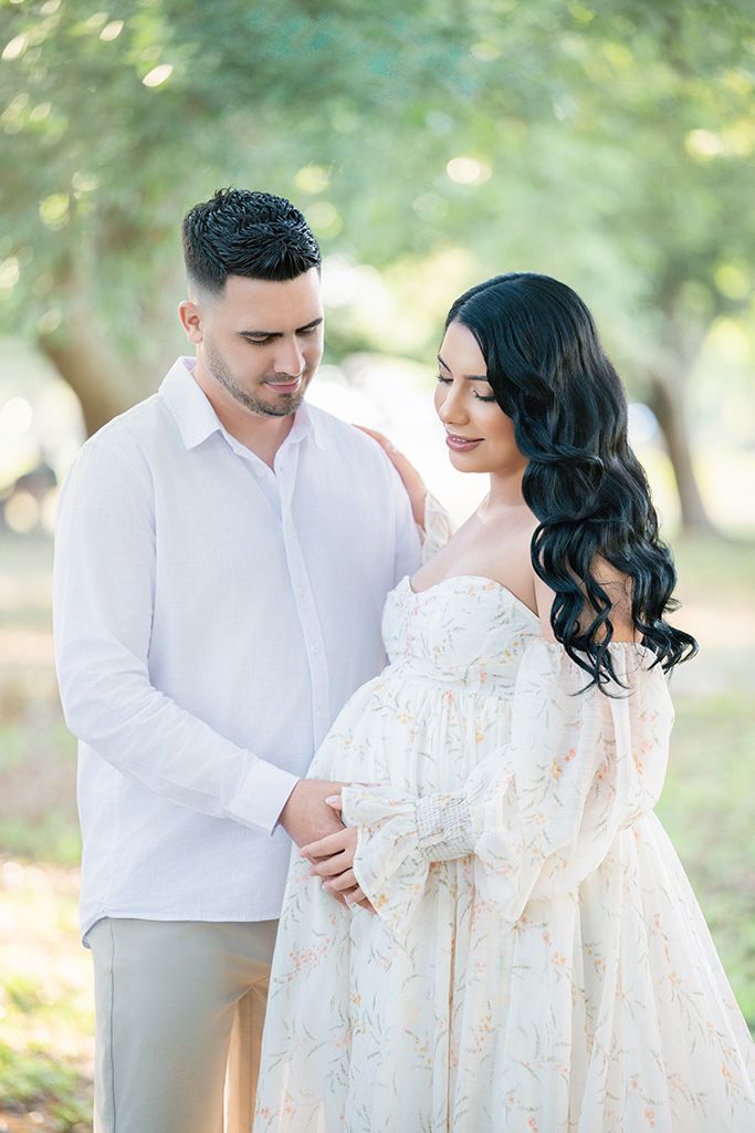 Expectant couple shares tender moment outdoors, man in white shirt gazes at womans baby bump in floral gown.
