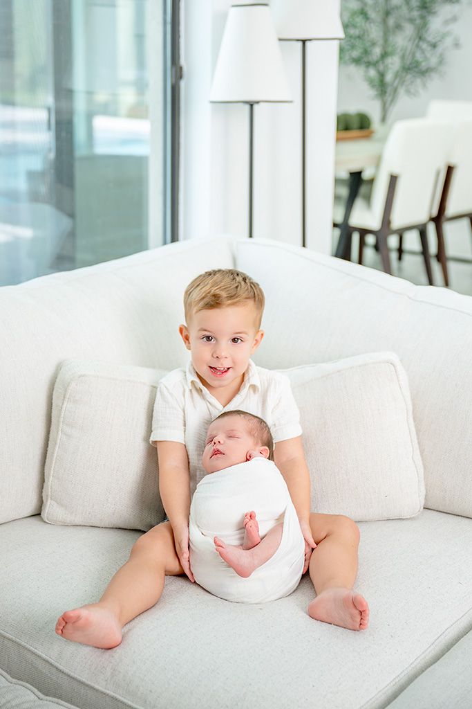 Young boy smiling on white sofa holding swaddled newborn baby sibling in bright modern living room.