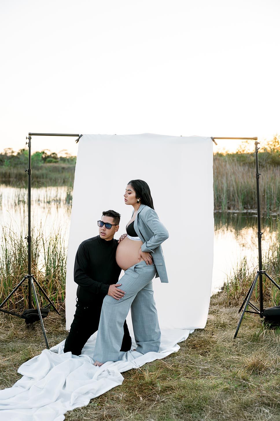 Couple posing for maternity photo shoot outdoors with white backdrop near lake at sunset.