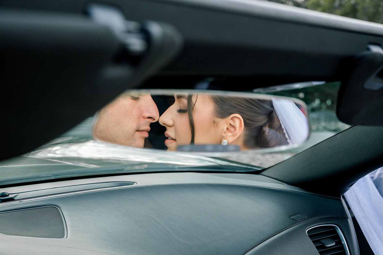 Bride and groom share a romantic kiss reflected in the rearview mirror of a luxury sports car.