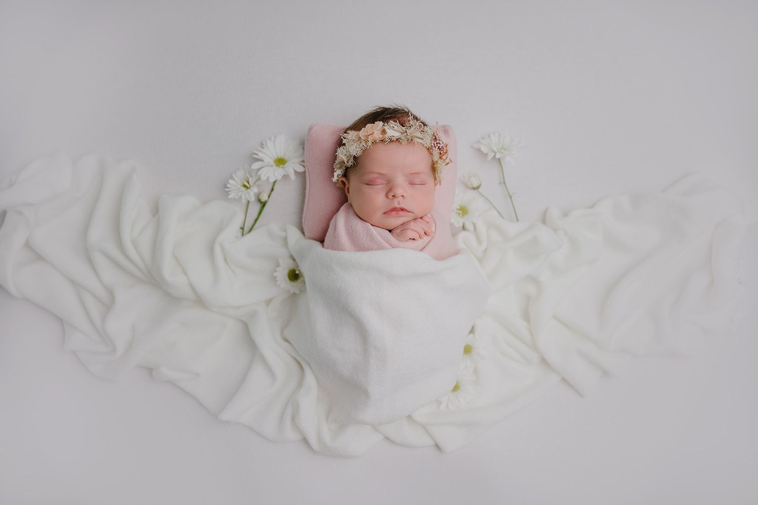 Newborn baby wrapped in white swaddle with floral crown, peacefully sleeping surrounded by white flowers on soft fabric.