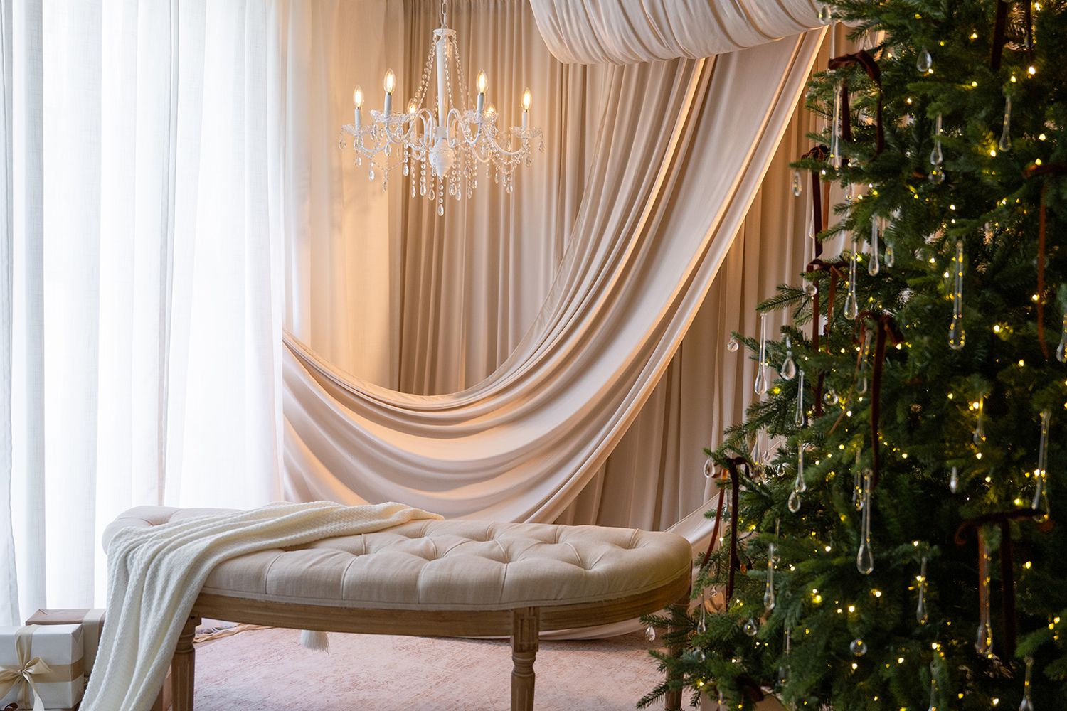 Elegant Christmas room with crystal chandelier, draped white curtains, tufted bench, and decorated Christmas tree with warm lights.