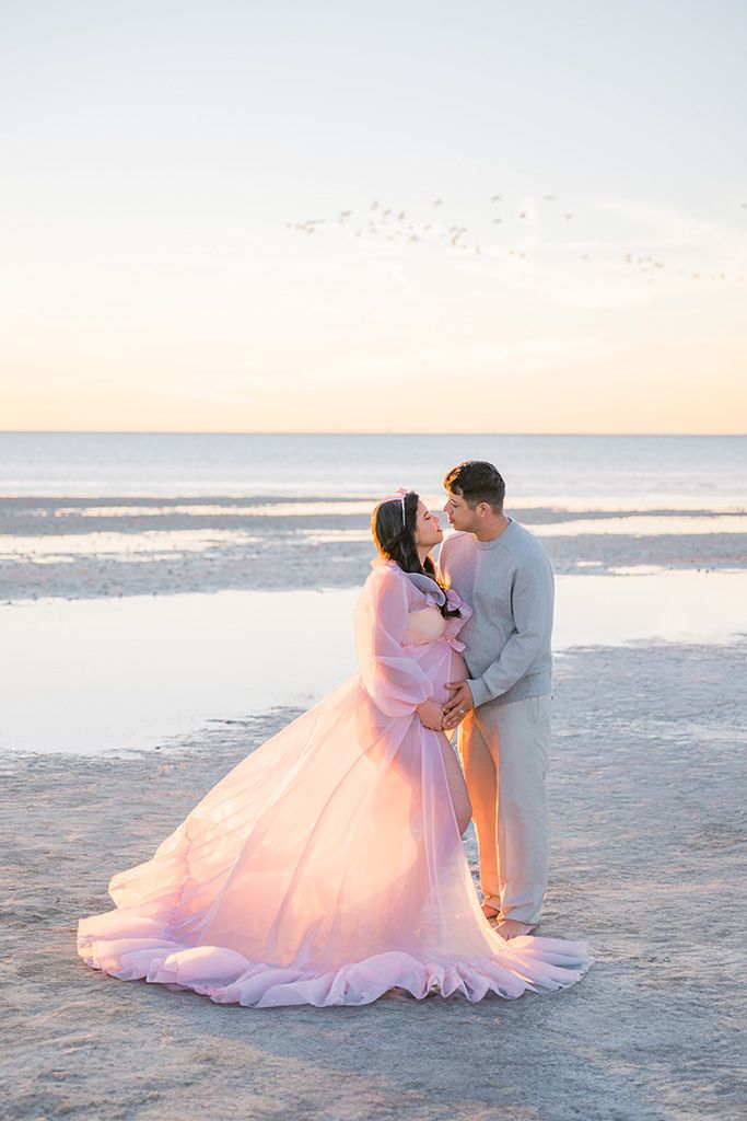 Couple embracing on beach at sunset, pregnant woman in flowing pink gown, man in gray, ocean backdrop.