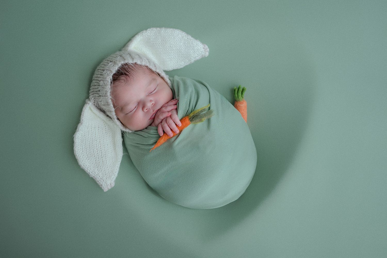 Newborn baby sleeping in green wrap with bunny ear hat holding a tiny carrot prop on green background.