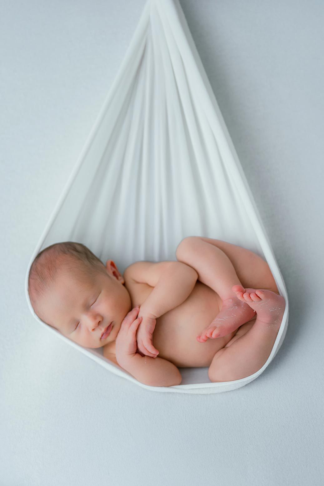 Newborn baby sleeping peacefully in a white fabric hammock, curled in fetal position on light blue background.