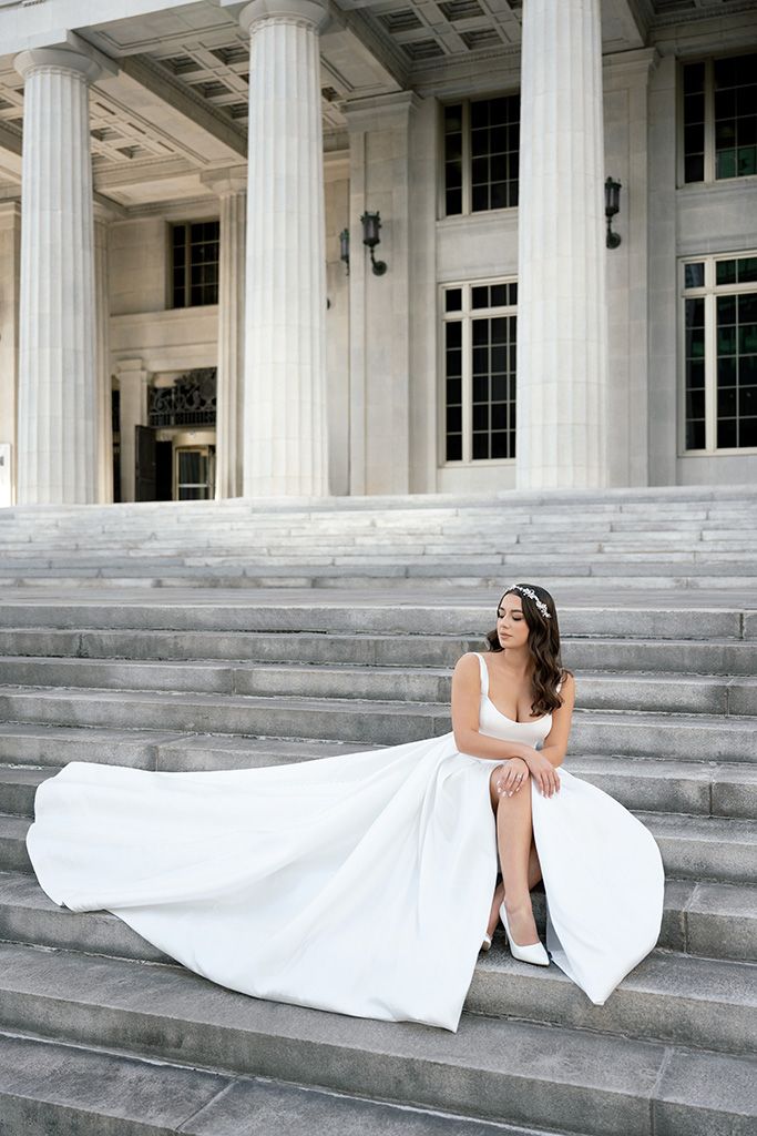 Bride in white gown with flowing skirt sits gracefully on stone steps of a neoclassical building with tall columns.