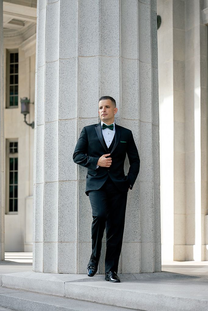Groom in black tuxedo with green bow tie leans against stone column at classical architecture venue.