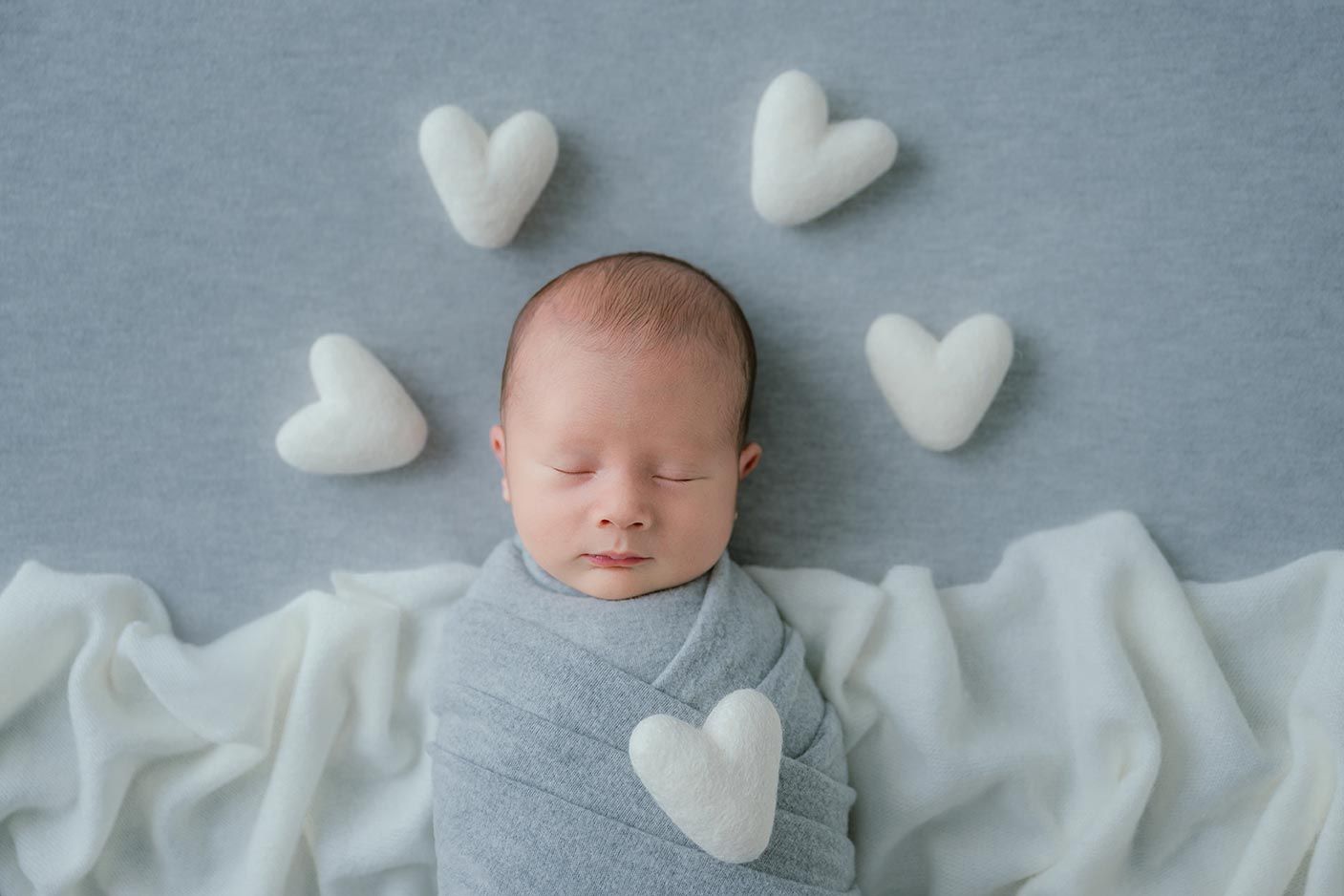 Newborn baby swaddled in gray wrap sleeping peacefully surrounded by white heart-shaped props on a soft blue background.