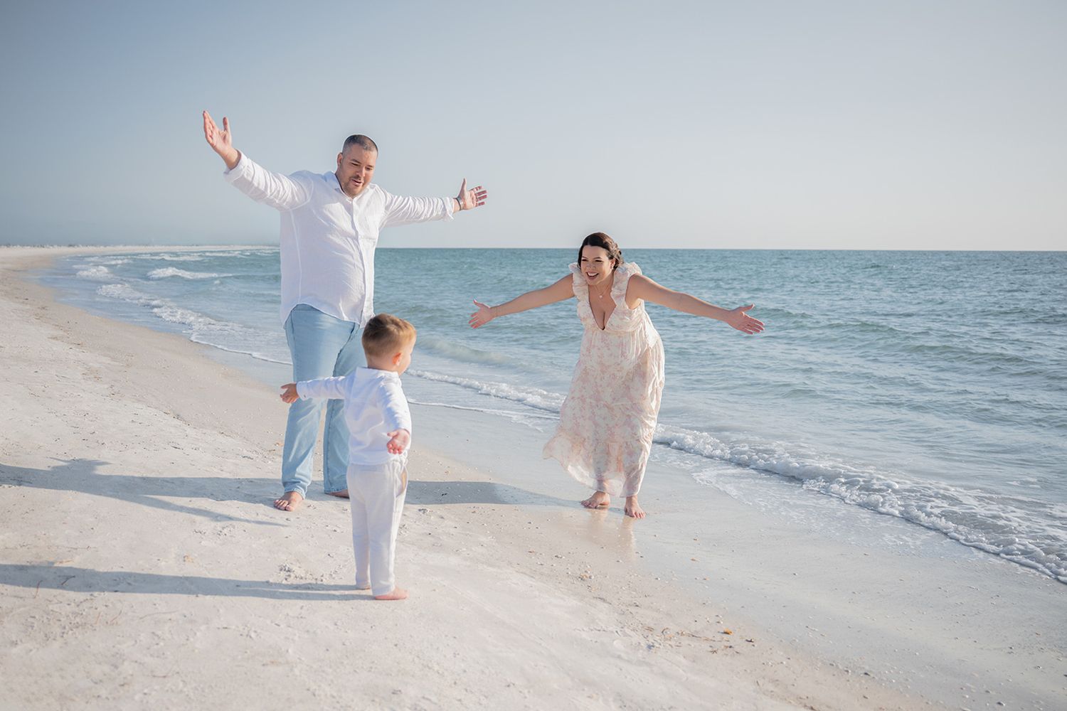 Family of three celebrating on a white sand beach, parents with arms raised joyfully as toddler runs toward them.