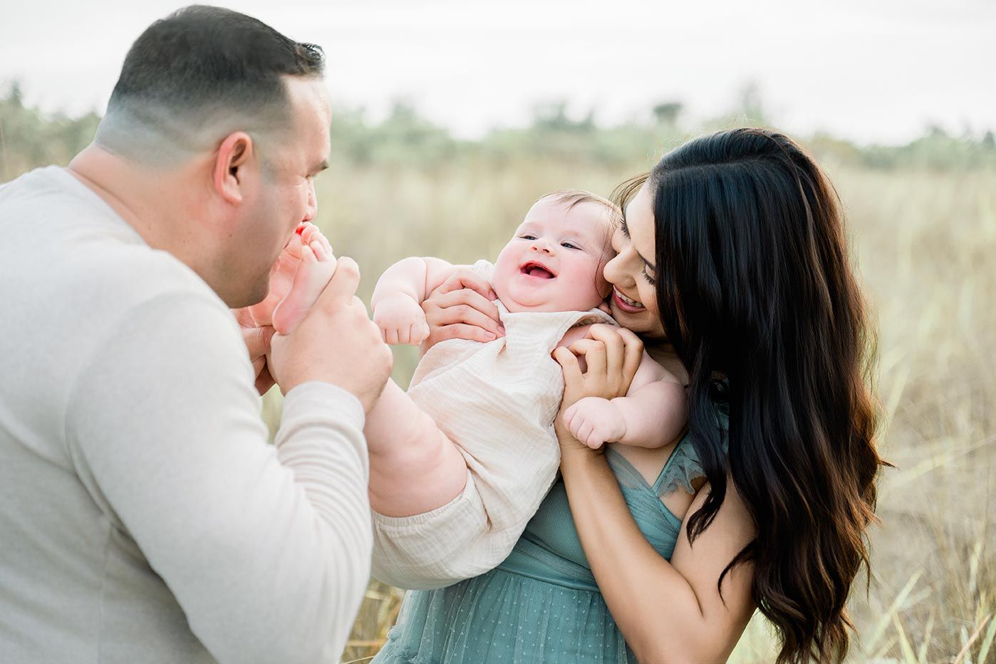 A joyful family photo with parents holding and kissing their laughing baby in a golden field.