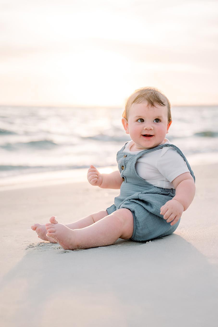 Adorable baby wearing teal blue overalls sits on sandy beach at sunset creating sweet candid moments.