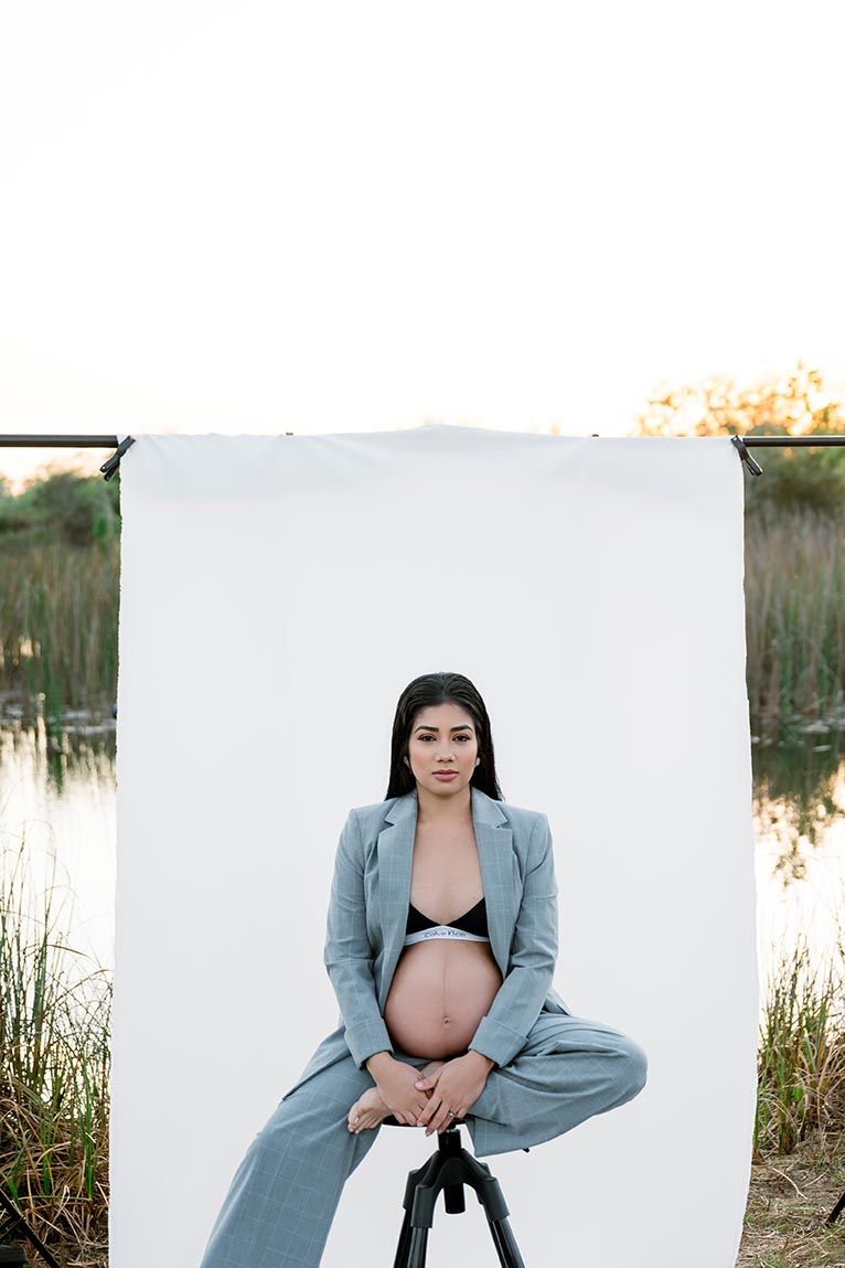 Pregnant woman in gray suit sits on tripod outdoors, posing against white backdrop near a serene lake.
