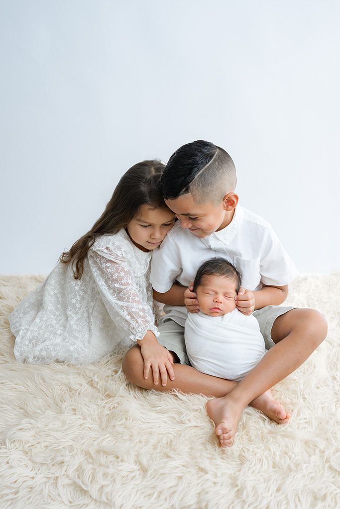 Two older siblings lovingly hold their sleeping newborn baby wrapped in white on a fluffy cream rug.