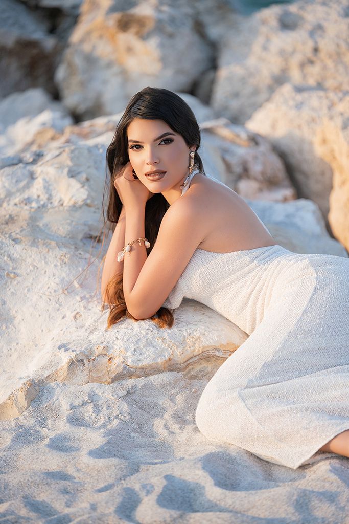 Elegant woman in white strapless dress posing on sandy rocks at golden hour beach.