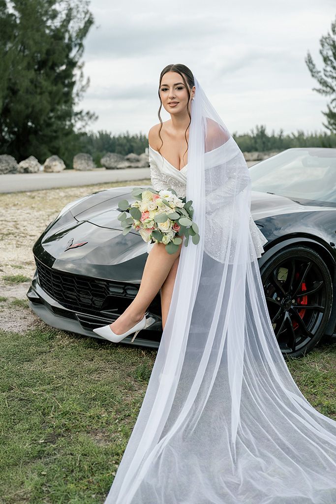 Bride in white wedding gown and veil poses with bouquet leaning against black Corvette outdoors.