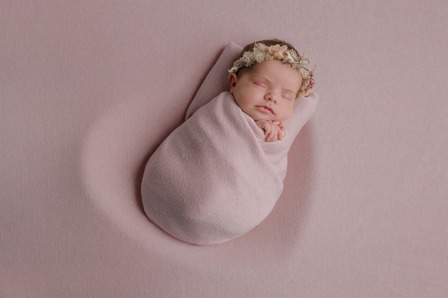 Newborn baby girl swaddled in pink wrap wearing floral crown, sleeping peacefully on pink backdrop.