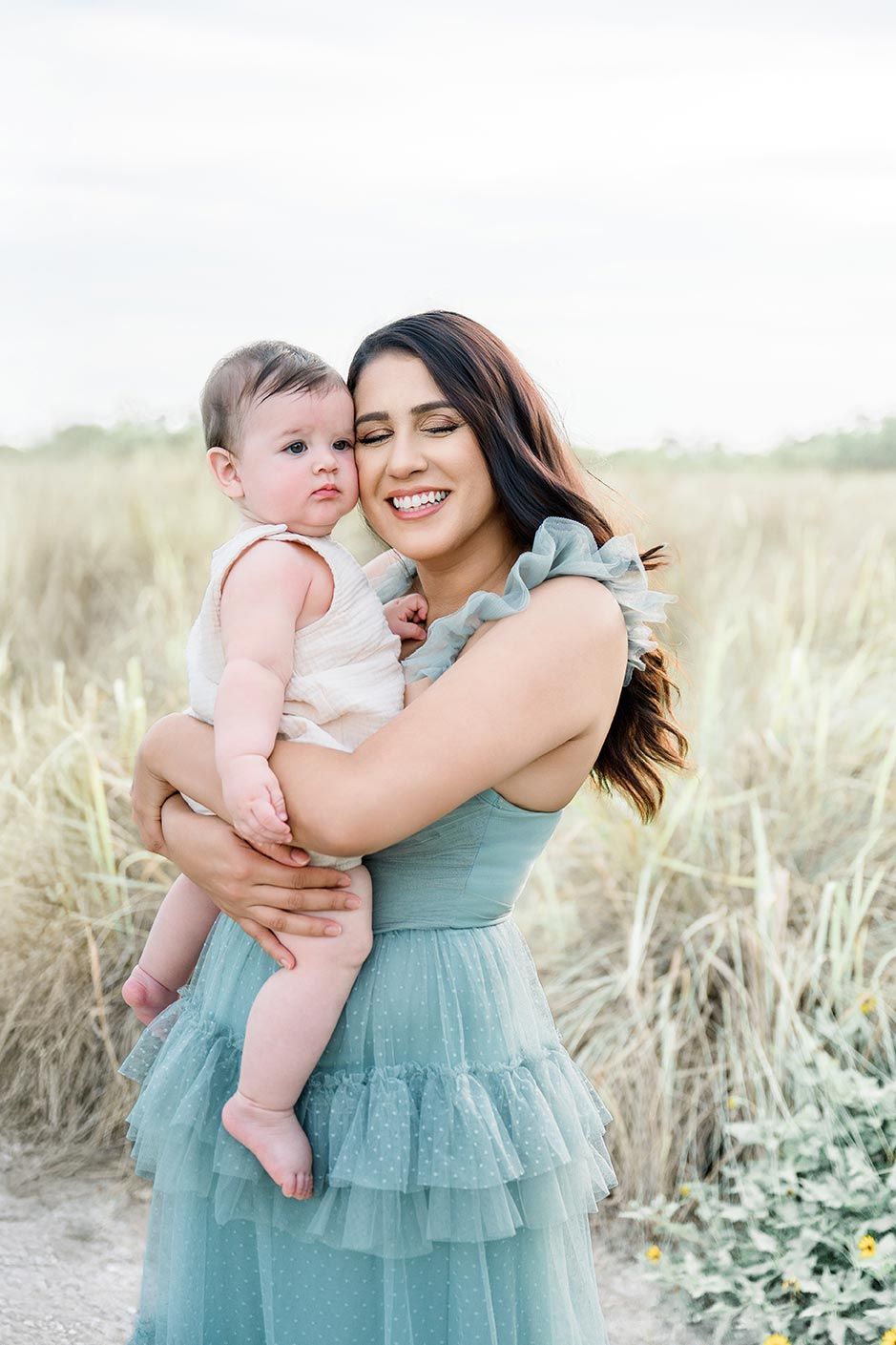 Mother and baby share tender moments in soft turquoise dress against natural beach grass setting.