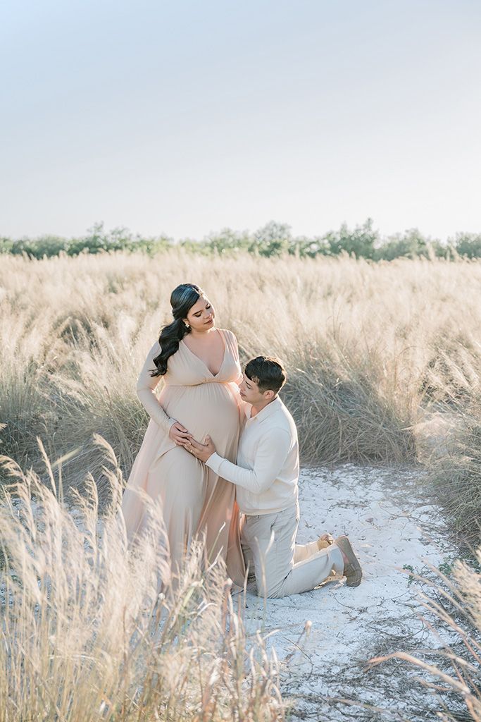 Couple in beige outfits pose in golden grass field, pregnant woman in flowing gown with partner kneeling beside her.